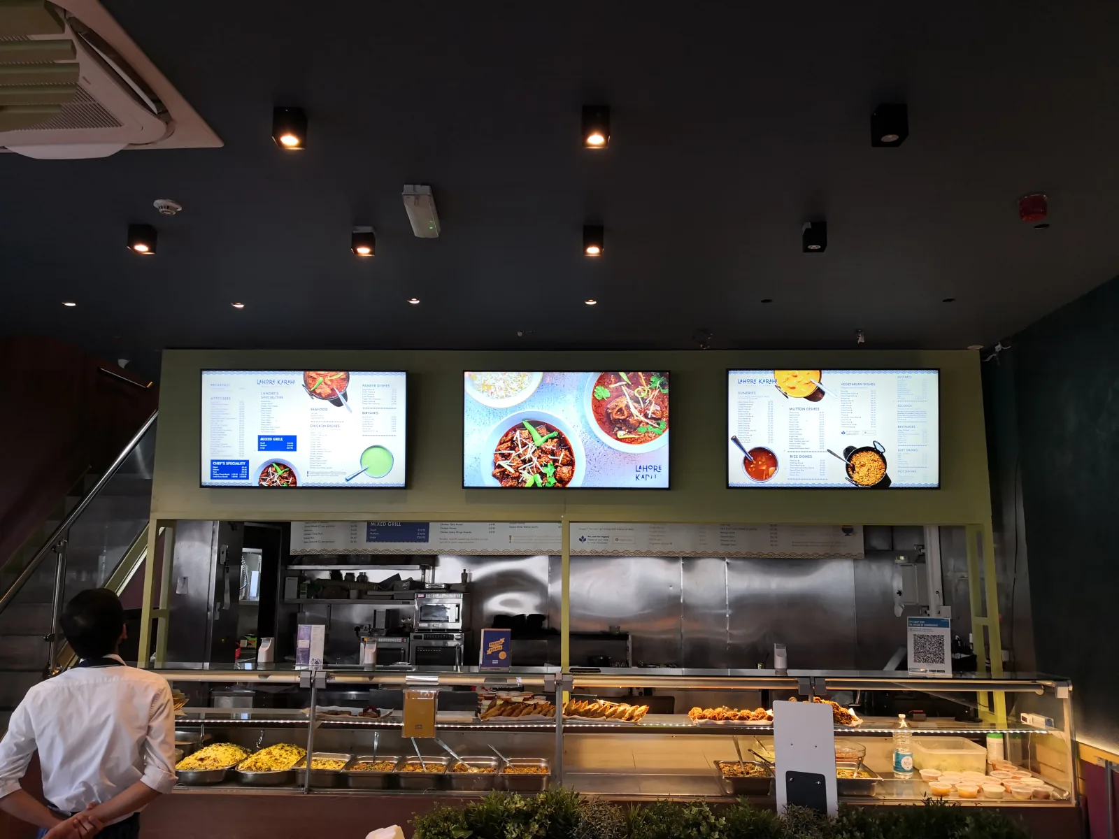 Three digital menu boards above a restaurant food counter